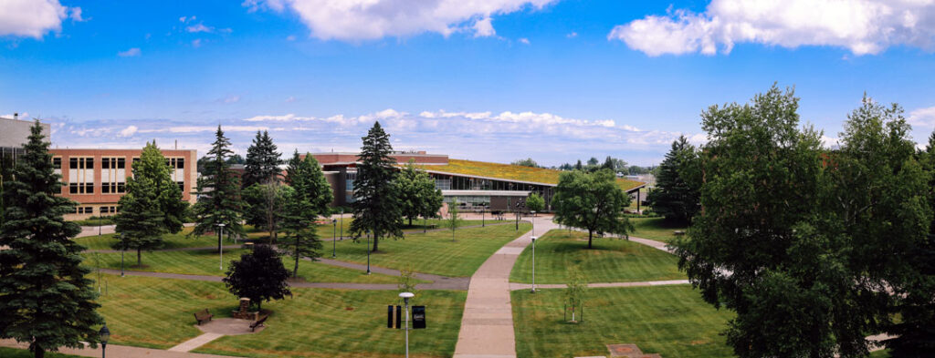 A panoramic view of a university campus with green lawns, trees, walkways, and modern academic buildings under a blue sky with scattered clouds welcomes students starting their freshman application.