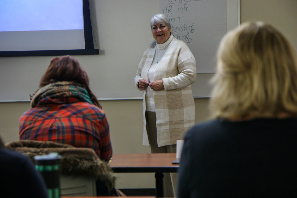 An educator stands and smiles while speaking to a small group in a classroom. She is in front of a whiteboard, and several people are seated at desks facing her.