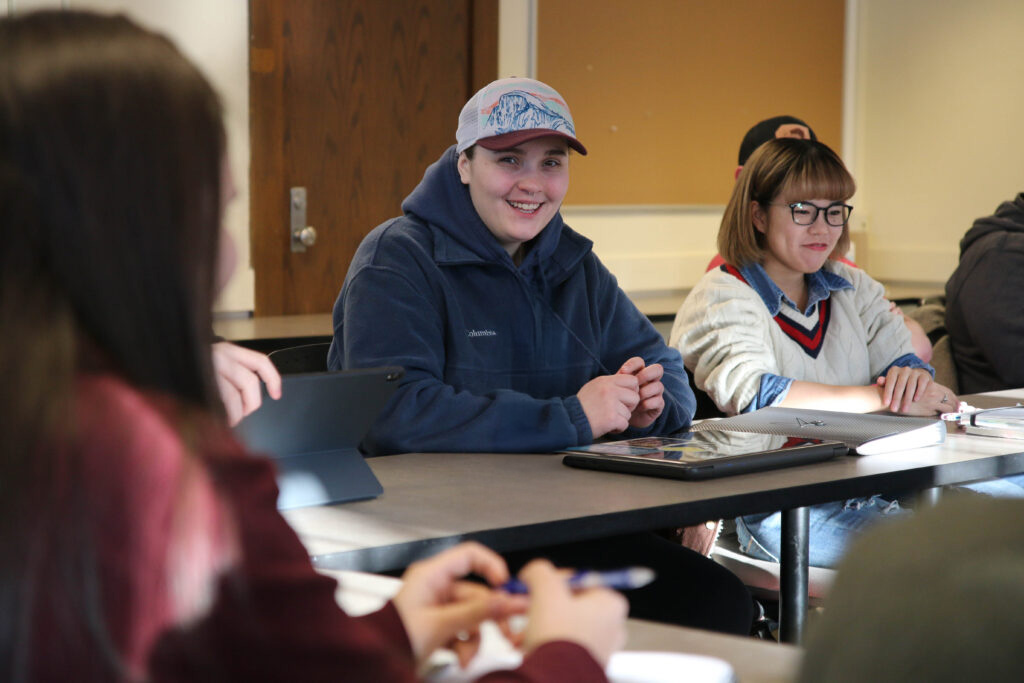 Several students sit around a table in a classroom, engaged in discussion. One student in a blue jacket and cap is smiling at the camera, while others listen and take notes.