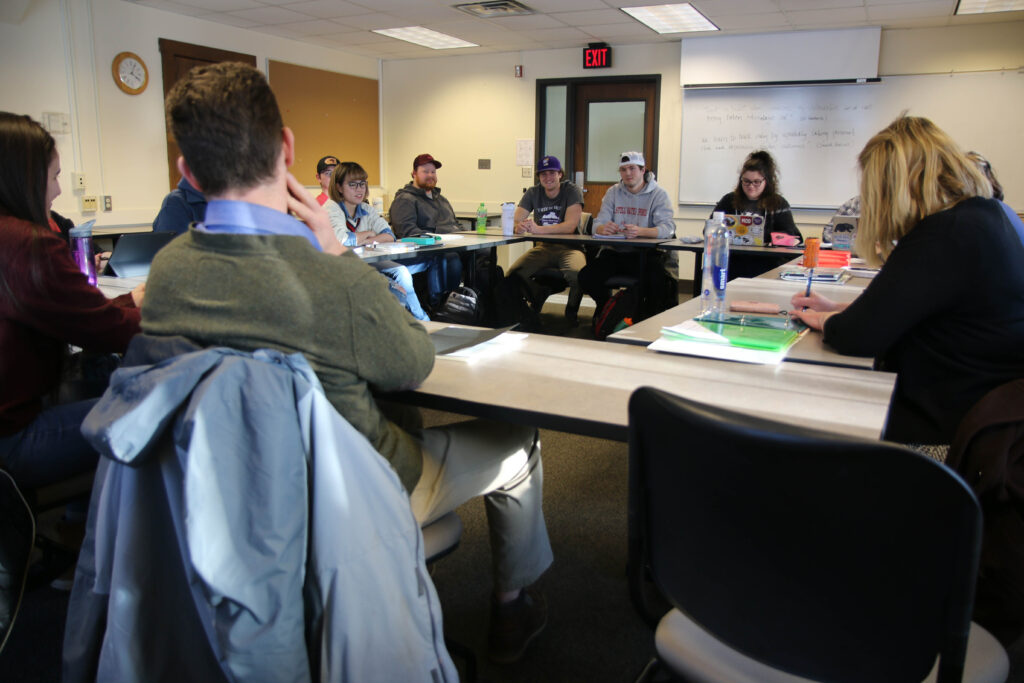 Students and a teacher sit around a large table in a classroom, engaged in discussion. Laptops, notebooks, and water bottles are on the tables, and a whiteboard with writing is visible at the front of the room.