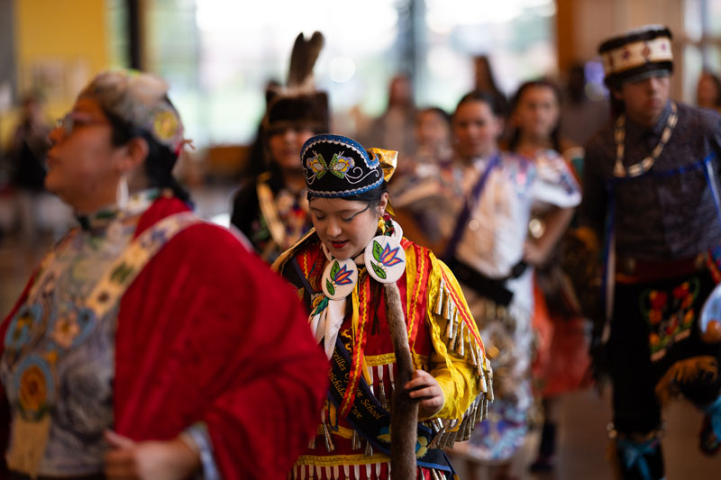 A group of people in vibrant traditional Indigenous regalia participate in a multicultural initiatives event indoors. The central figure wears a colorful headdress and holds a decorated staff, with others in the background.