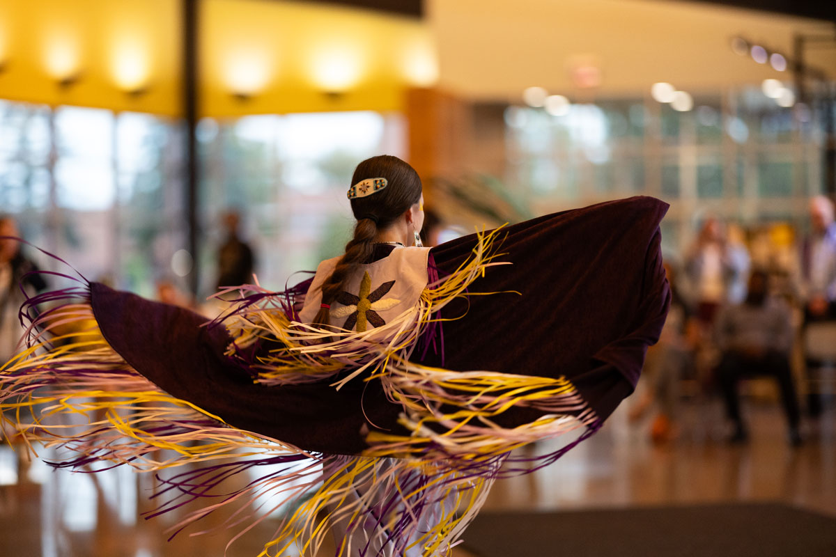 A person in traditional regalia dances indoors, purple, yellow, and white fringe swirling as they spin. The scene highlights community and the importance of Education Law Ethics and Communication in Schools, with onlookers near sunlit windows.