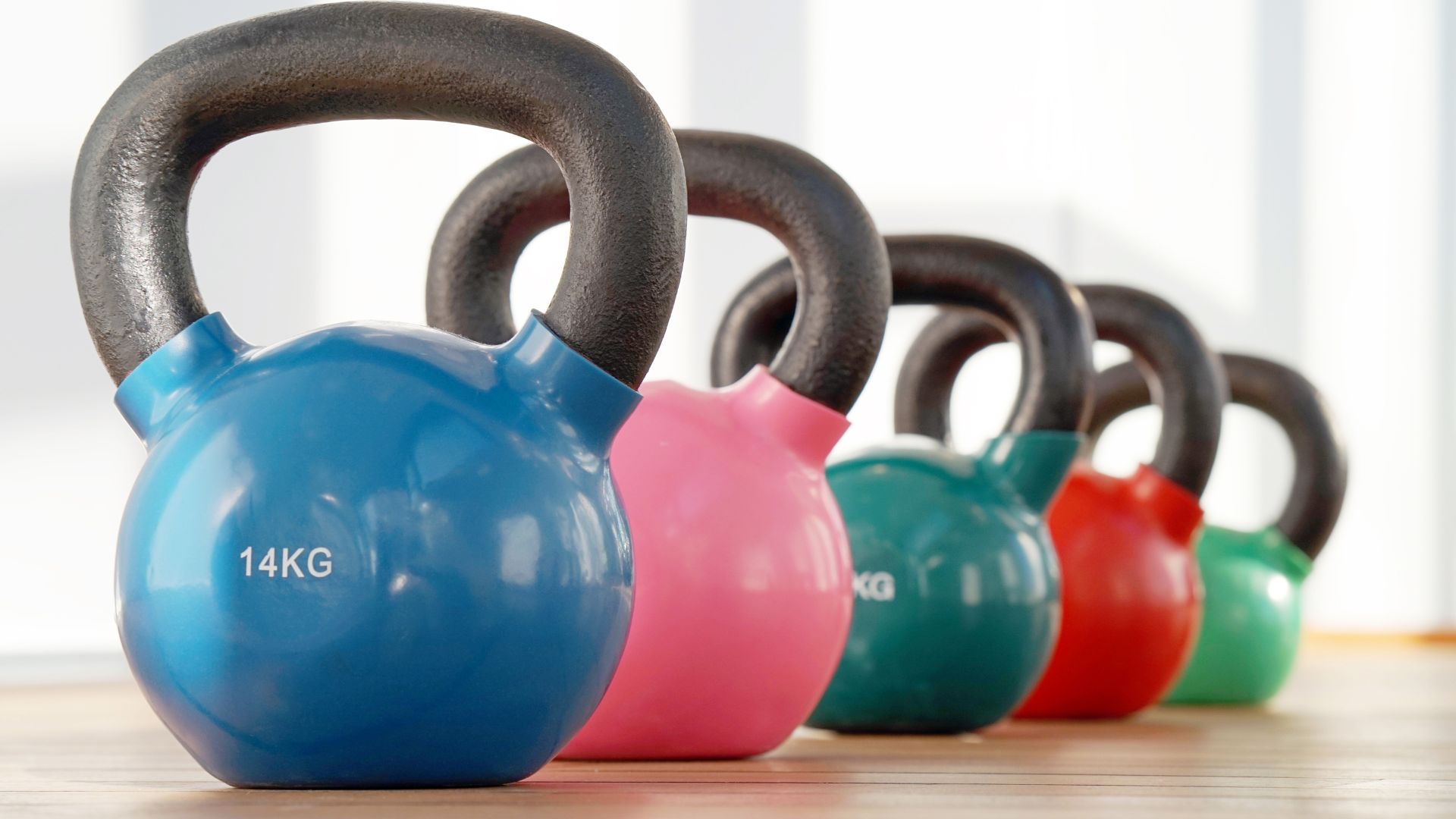 A row of colorful kettlebells, including blue, pink, green, and red, is lined up on a wooden floor. The blue kettlebell in front is labeled 14 KG. Bright, natural light fills the background.