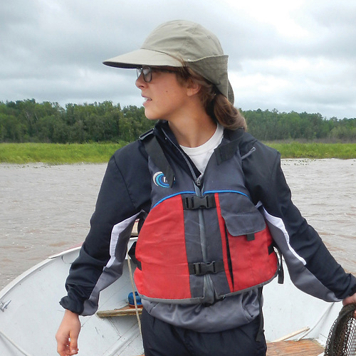 A person wearing glasses, a gray hat, and a red life jacket stands on a boat, looking to the side. The background shows a river and trees under a cloudy sky.