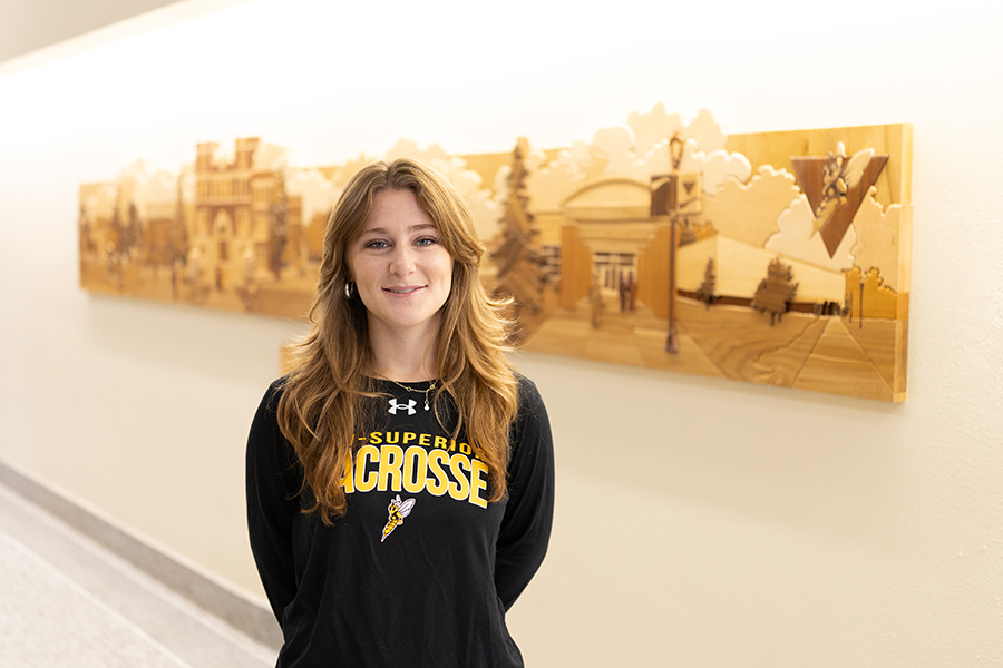 A woman wearing a black U-Superior Lacrosse long-sleeve shirt stands in front of a wooden relief.