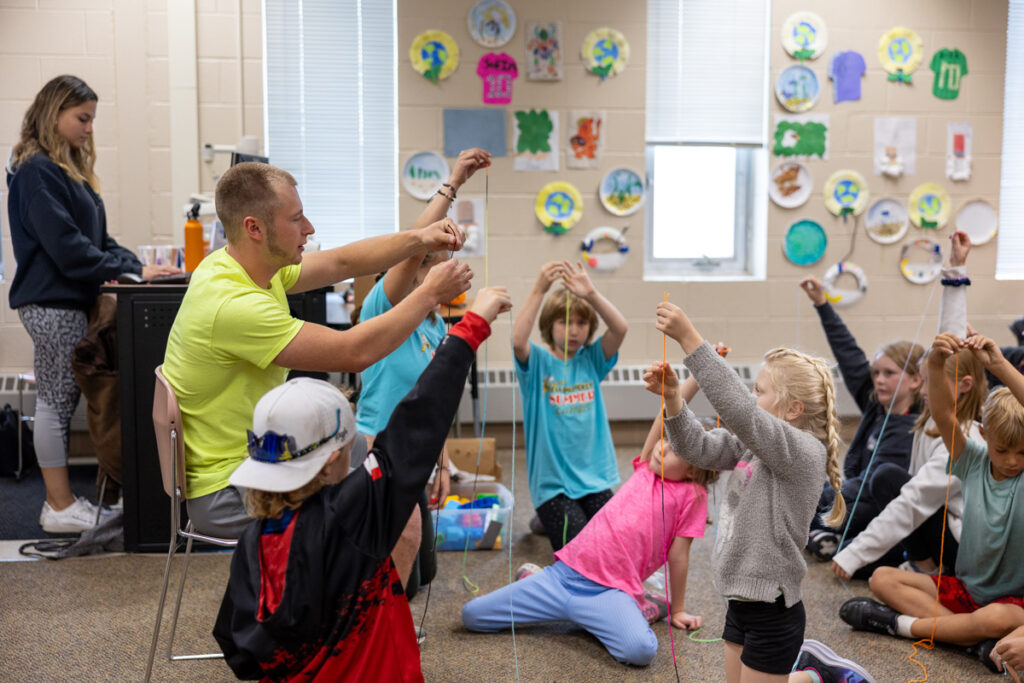 An adult sits and holds up string while a group of children kneel or sit around him, each raising strings—typical of hands-on activities at Summer Camp. A woman works at a computer in the background; colorful art projects line the classroom wall.