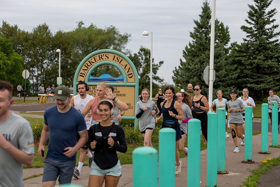 People jogging past a Barker’s Island sign on a cloudy day with trees in the background.