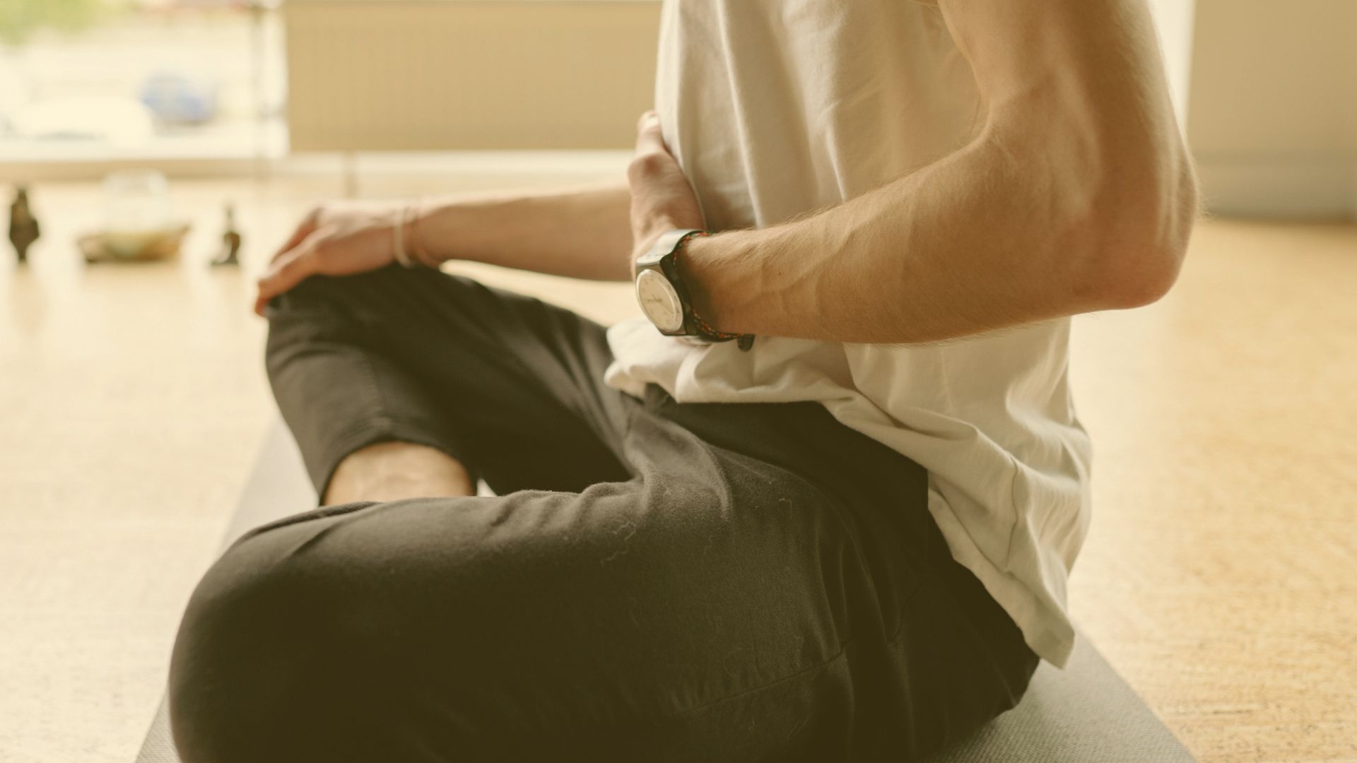 A person sitting cross-legged on a mat indoors, wearing a white shirt and black pants, with one hand resting on their abdomen, possibly practicing meditation or breathing exercises.