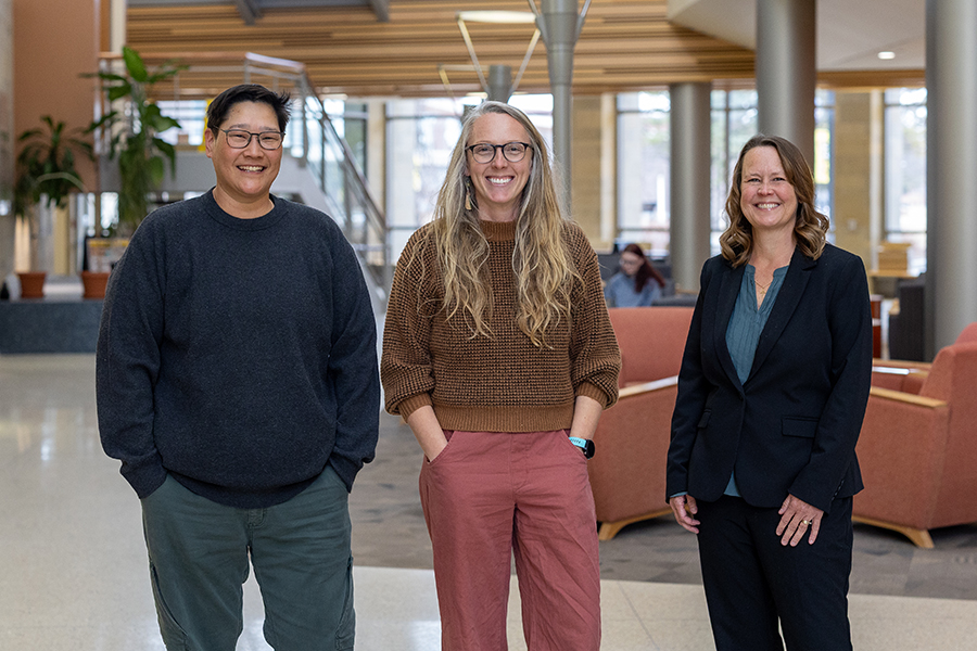 Three people stand indoors, smiling at the camera. Two are casually dressed in sweaters and pants, while one wears a dark suit. They are in a bright, modern lobby with plants, large windows, and comfortable seating.