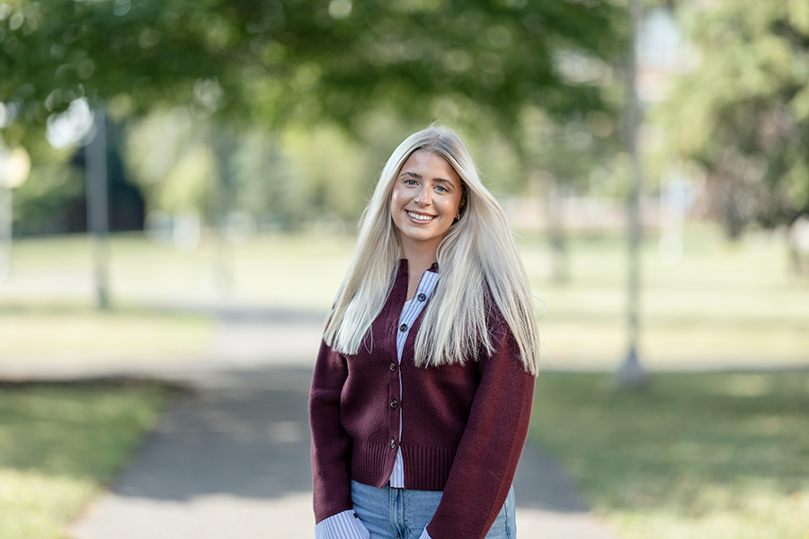 Woman with long blonde hair stands outdoors on a path, wearing a maroon sweater and blue jeans.
