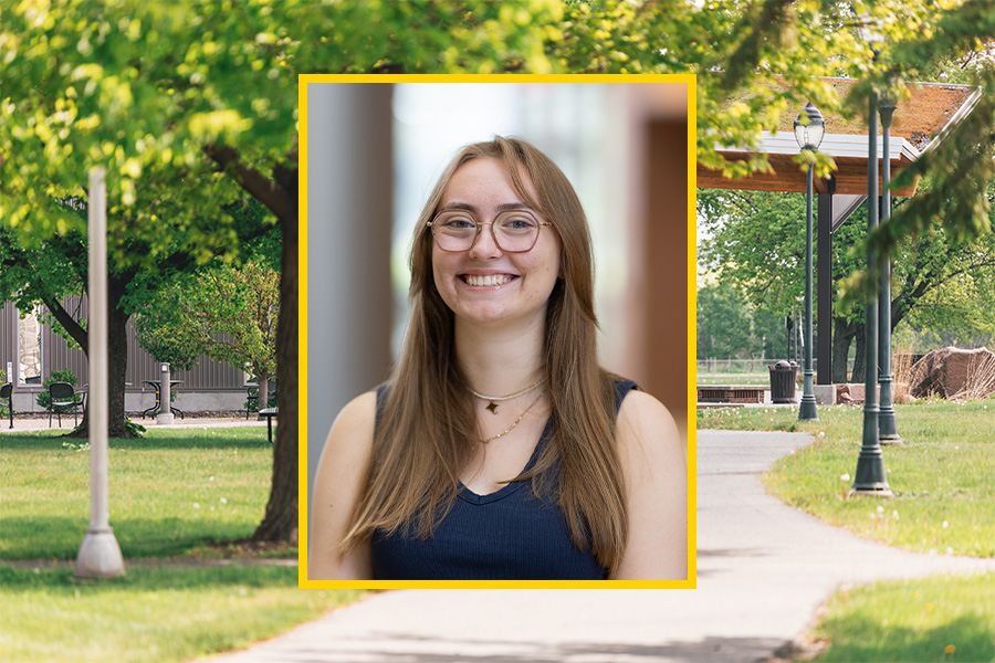 A young woman with long brown hair and glasses smiles, standing outdoors in front of a blurred background of trees and a park path. The image is framed with a bright yellow border.