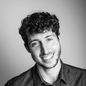 A man with short curly hair and a beard smiles at the camera, perhaps recalling past events and activities. He is wearing a button-up shirt and is photographed in black and white against a plain background.