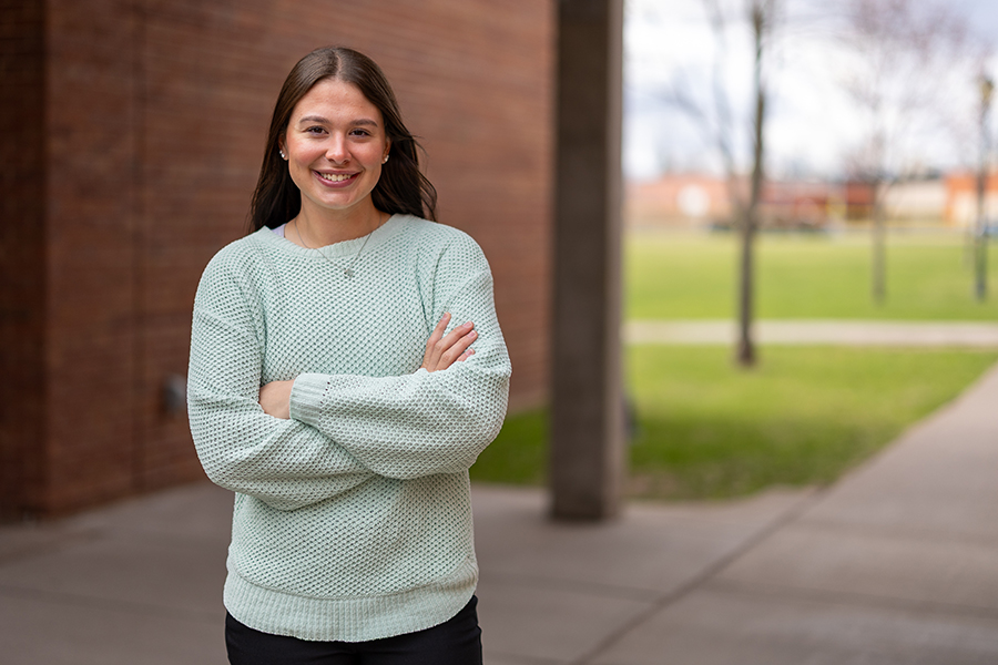 Woman wearing a light green sweater smiles with arms crossed outside near a brick building.