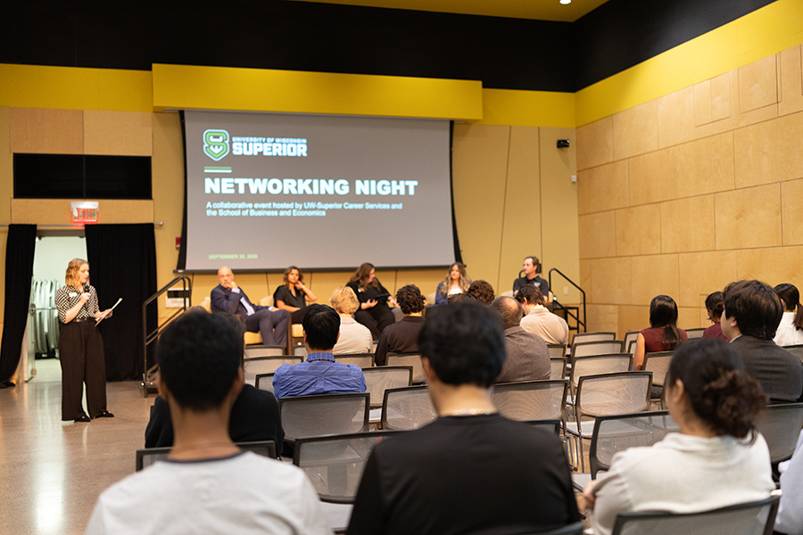 A woman speaks at a networking event with panelists seated onstage and audience listening.