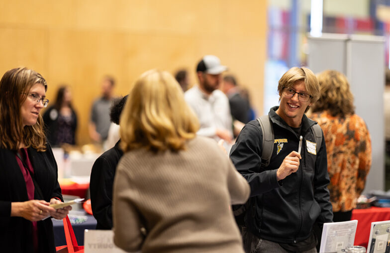 People talking and networking at an indoor event with tables and informational displays.