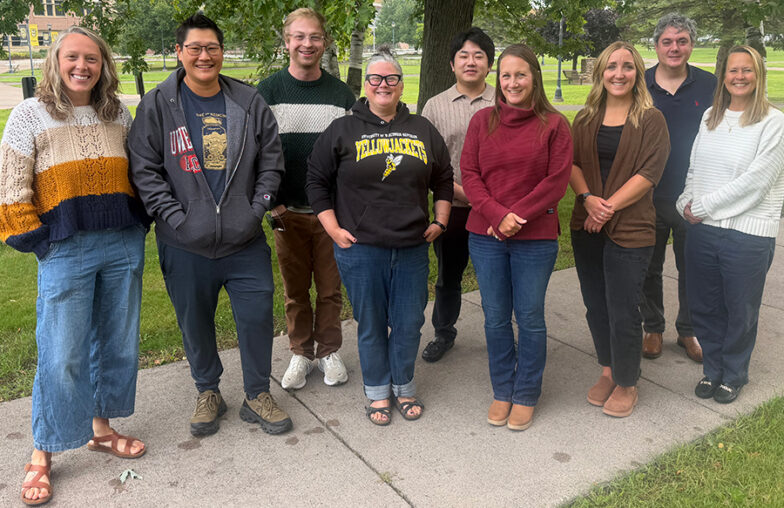 Eight people stand in a row on a sidewalk outdoors.