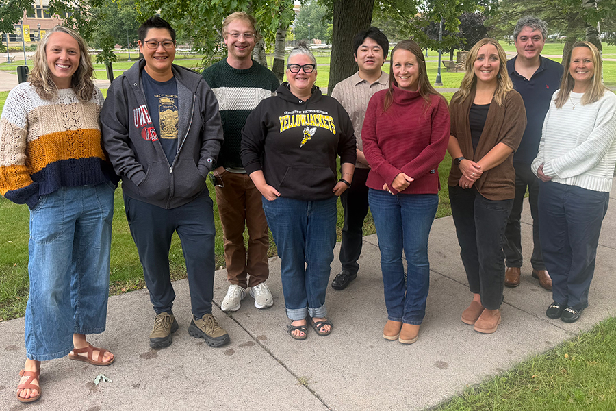 Eight people stand in a row on a sidewalk outdoors.