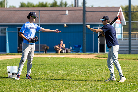 Two people practice baseball on a field, one tossing a ball and the other holding a bat.