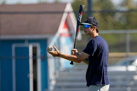 Person holding a baseball and bat standing outdoors near a blue building and bleachers.