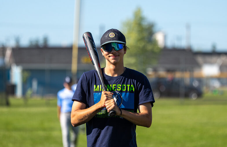 Person wearing sunglasses and a Minnesota shirt holds a baseball bat on a grassy field.