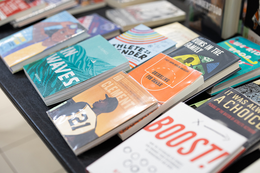 A display of various books arranged on a table, featuring colorful covers and diverse titles, including “In Waves,” “BOOST!,” “It Was All a Hoax,” and several others overlapping each other.