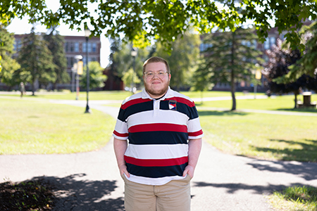 Person standing on a sunlit campus path, wearing a striped shirt and khaki shorts.