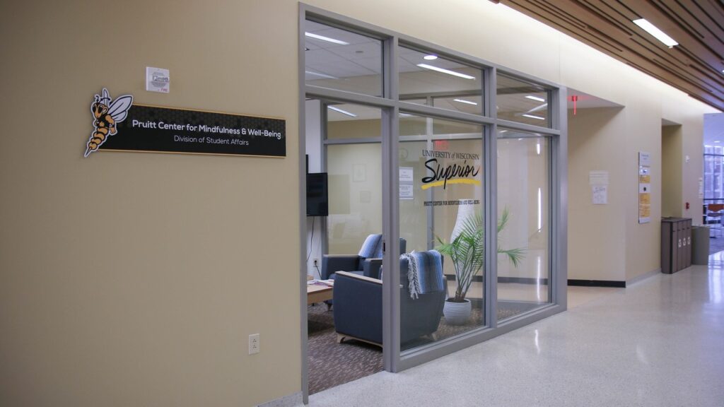 A glass-walled office labeled Pruitt Center for Mindfulness & Well-Being with cozy seating, plants, and a TV inside. A sign with a yellowjacket mascot is mounted beside the door.
