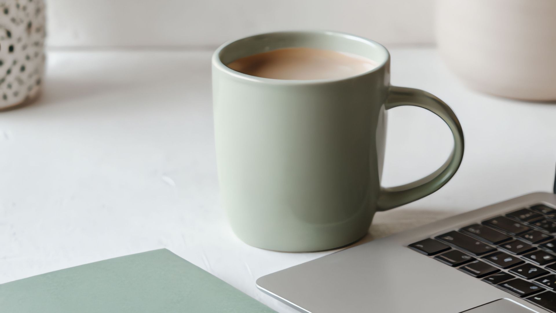 A green ceramic mug filled with a light brown beverage sits next to a partially visible laptop on a white desk, with a notebook and a decorative item in the background.