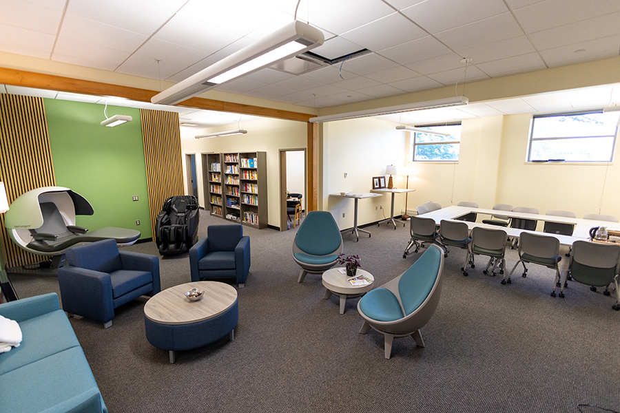 Modern lounge with blue chairs, round tables, a bookshelf, and conference table in the back.