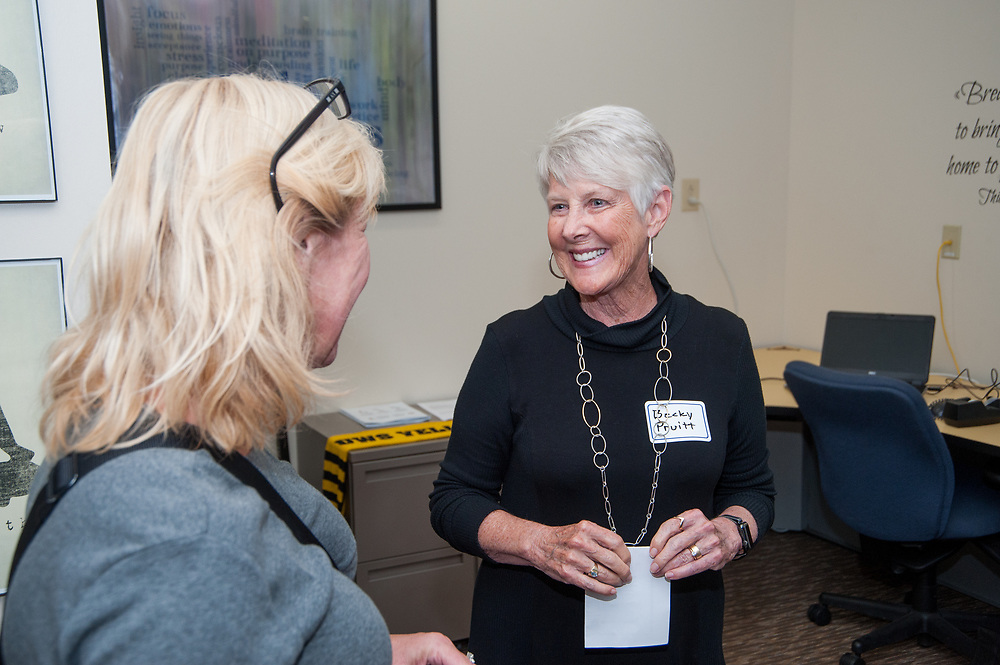 Two women converse in an office. One, with short white hair and a name tag from the Pruitt Center for Mindfulness and Well-Being, smiles warmly as she holds a white envelope. The other woman, blonde and wearing glasses, listens attentively.