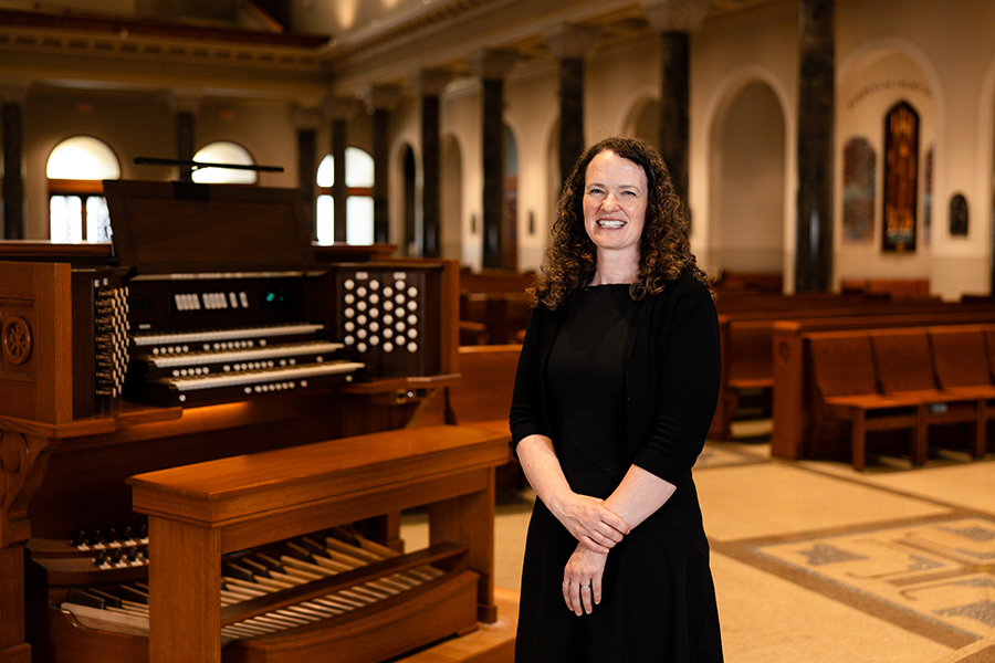 A woman in a black dress stands beside a pipe organ in a church.