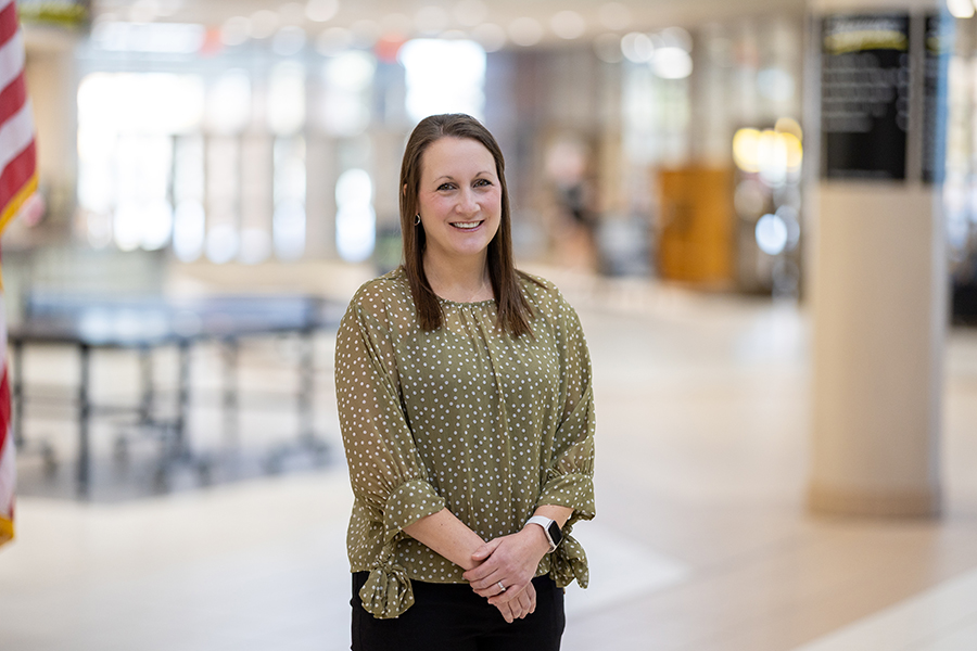 A woman with straight brown hair, wearing a green blouse with white polka dots and black pants, stands smiling in a bright, spacious indoor setting with blurred background elements.
