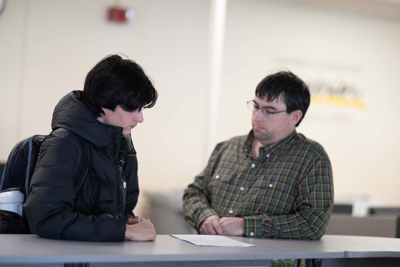Two people sit across from each other at a table, engaged in conversation. One is wearing a black puffer jacket, while the other wears glasses and a plaid shirt. A paper with academics notes lies on the table between them.