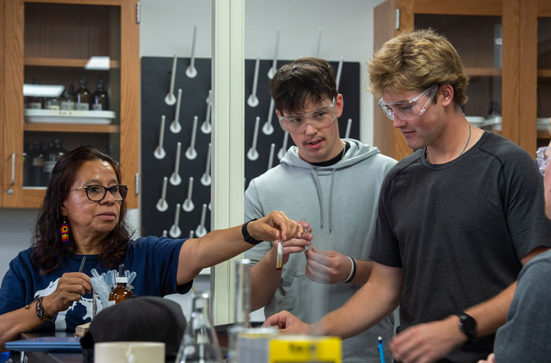 A woman wearing safety glasses shows a test tube to two students, also in safety glasses, in a UW-Superior science lab with glassware and shelves in the background.