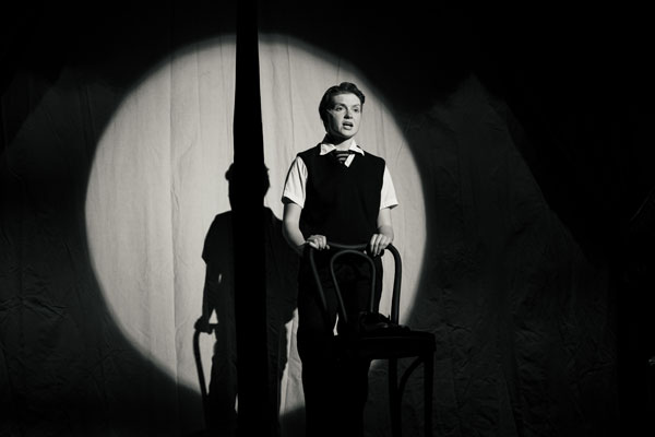 A filmmaking scene: person by chair in a spotlight, casting shadow on curtain. Black and white.