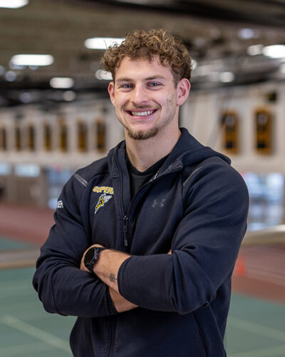 A man with curly hair stands smiling, arms crossed, in an indoor athletic facility.