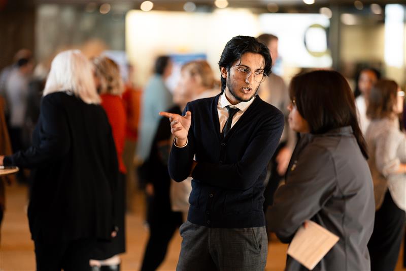 A man wearing glasses, a tie, and a dark cardigan gestures while discussing finance with a woman in a crowded indoor setting. Other people are mingling in the blurred background.