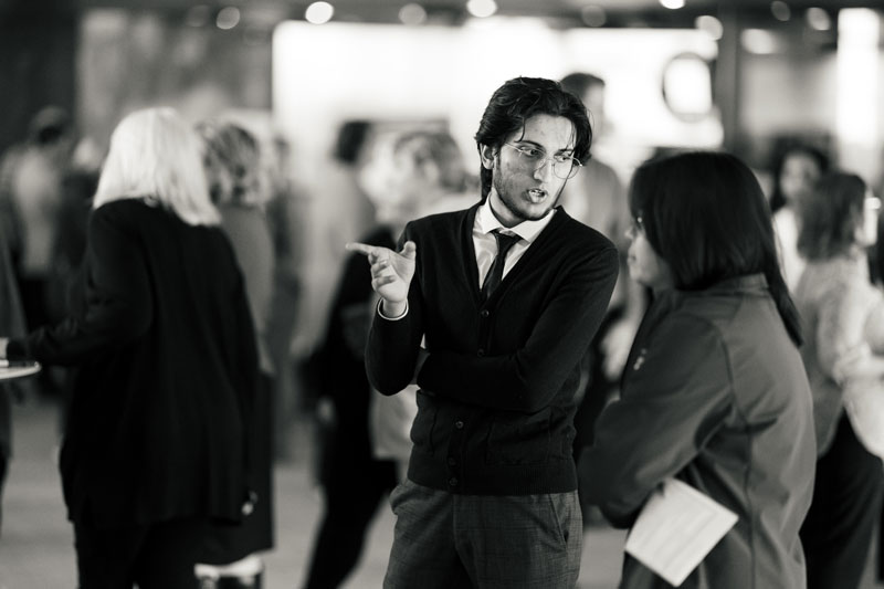 Man in glasses discusses finance while gesturing to a woman in a crowded indoor setting.