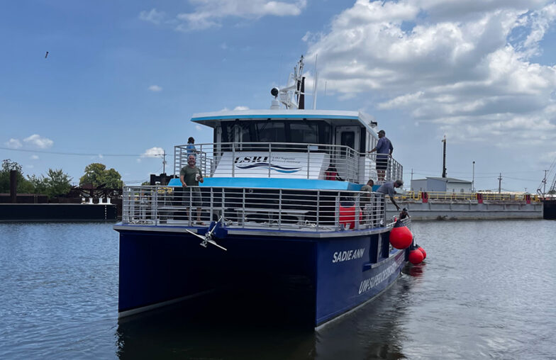 A blue research vessel named SAGE-ANN is docked with people on the deck under a partly cloudy sky.