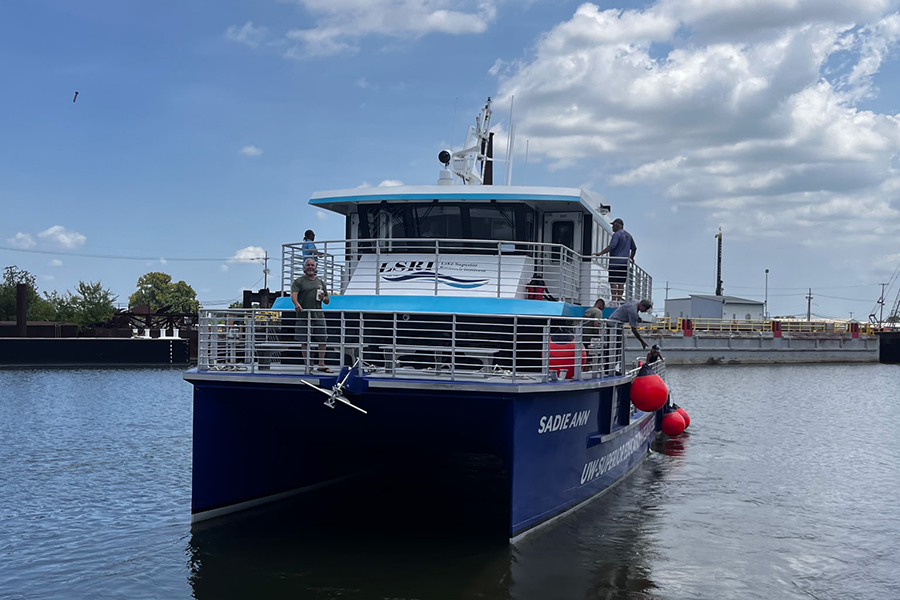 A blue research vessel named SAGE-ANN is docked with people on the deck under a partly cloudy sky.