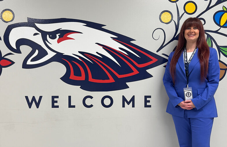 Woman in blue suit stands by a wall mural of an eagle head and the word WELCOME..
