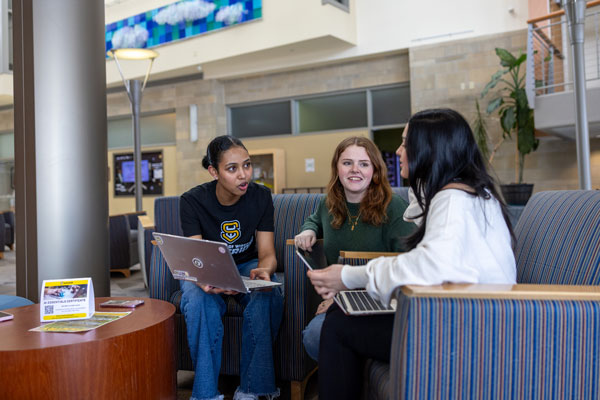 Three students sit on couches indoors, talking and using laptops around a small table.