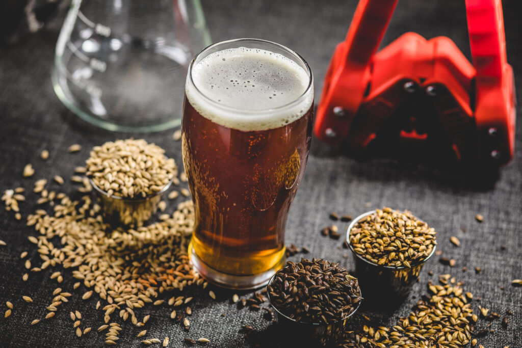 A pint glass of beer sits on a dark surface surrounded by small bowls of malted barley and grains. A red beer bottler and a glass flask are blurred in the background.