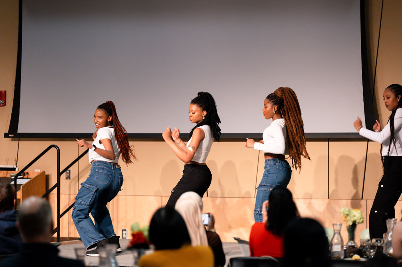 Four women are performing a dance on stage in front of an audience, showcasing Multicultural Initiatives. They wear white tops and blue jeans, moving in coordinated poses against a large blank screen.