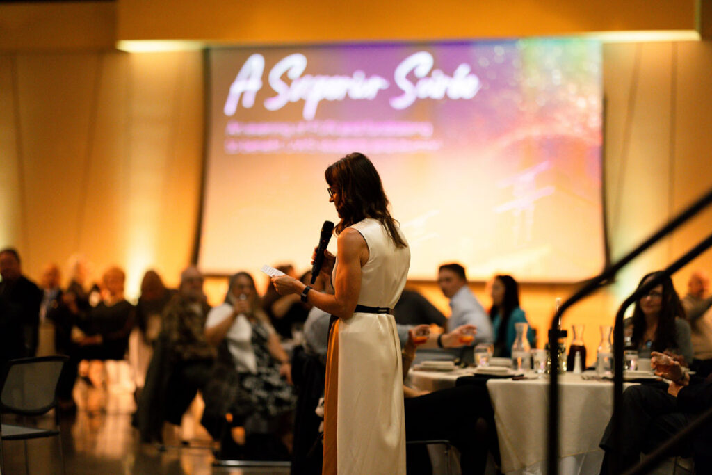 A woman discusses the Superior Fund at an indoor event with seated attendees and a projector screen.