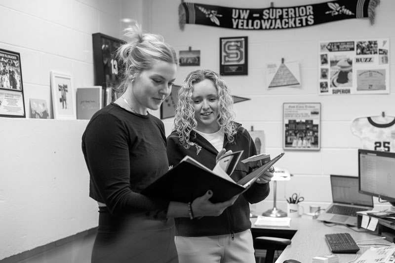 Two women in an office discuss notebooks; sports memorabilia shows Sport and Recreation Management.