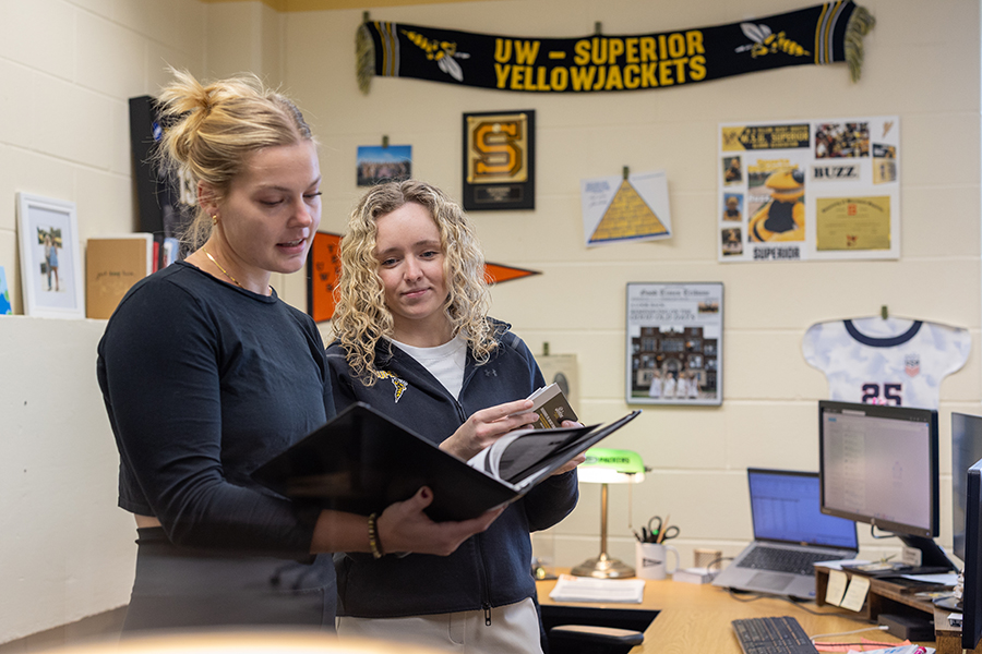 Two women stand in an office, looking at a binder together. The office walls are decorated with UW-Superior Yellowjackets memorabilia, framed photos, and a computer is on the desk nearby.