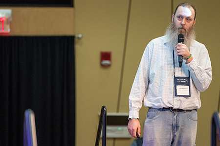 Man with a microphone speaks at an indoor event, wearing a conference badge and casual clothing.