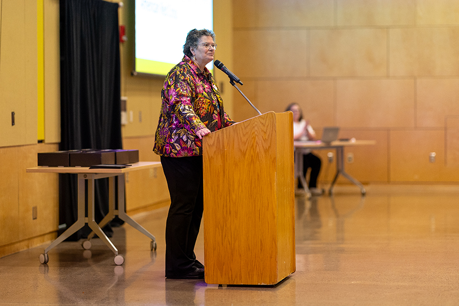 A person stands at a podium speaking into a microphone in a large indoor room.