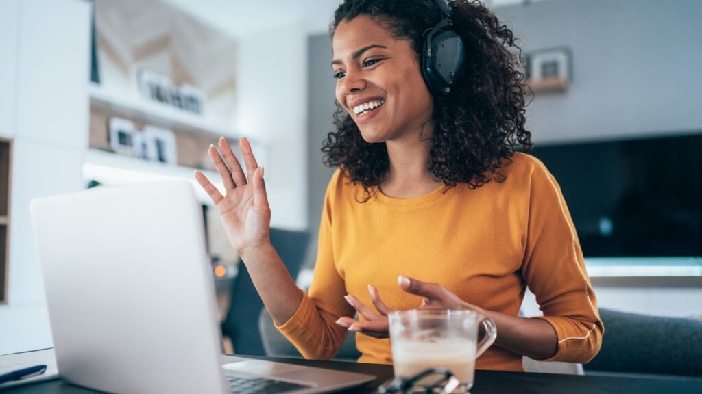 A woman wearing headphones and a yellow top smiles and waves at her laptop during a video call. A glass mug and eyeglasses sit on the table in front of her in a bright, modern room.